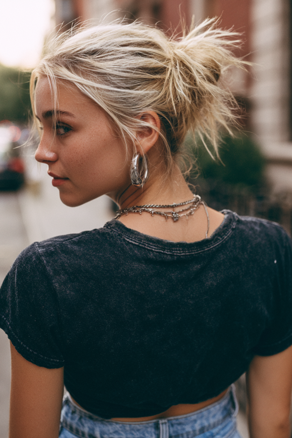 Portrait of a woman with a messy blonde bun, wearing hoop earrings and layered necklaces, in a casual black crop top outdoors