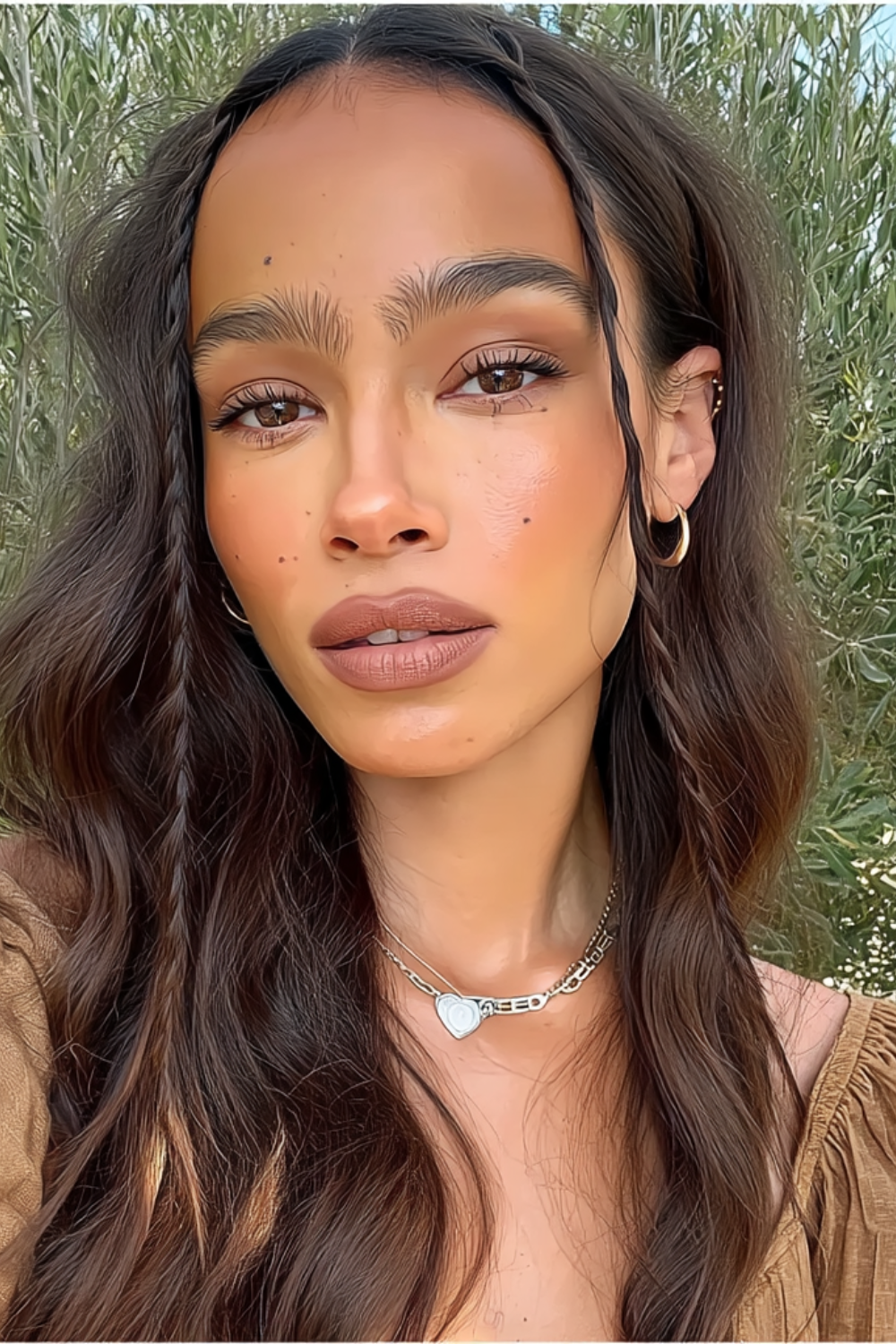 Close-up portrait of a woman with long brown hair, hoop earrings, and a heart necklace, outdoors with greenery in the background.