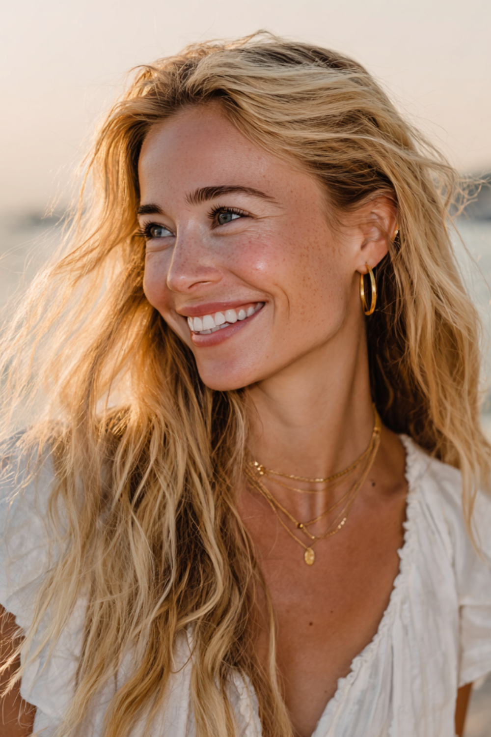 Close-up of a smiling blonde woman outdoors, wearing gold hoop earrings and layered gold necklaces.