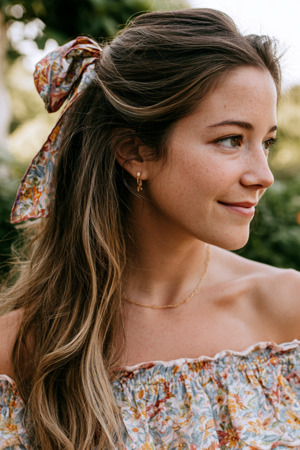 Close-up side profile of a smiling woman outdoors with a floral off-the-shoulder top, a patterned scarf in her hair, and gold earrings