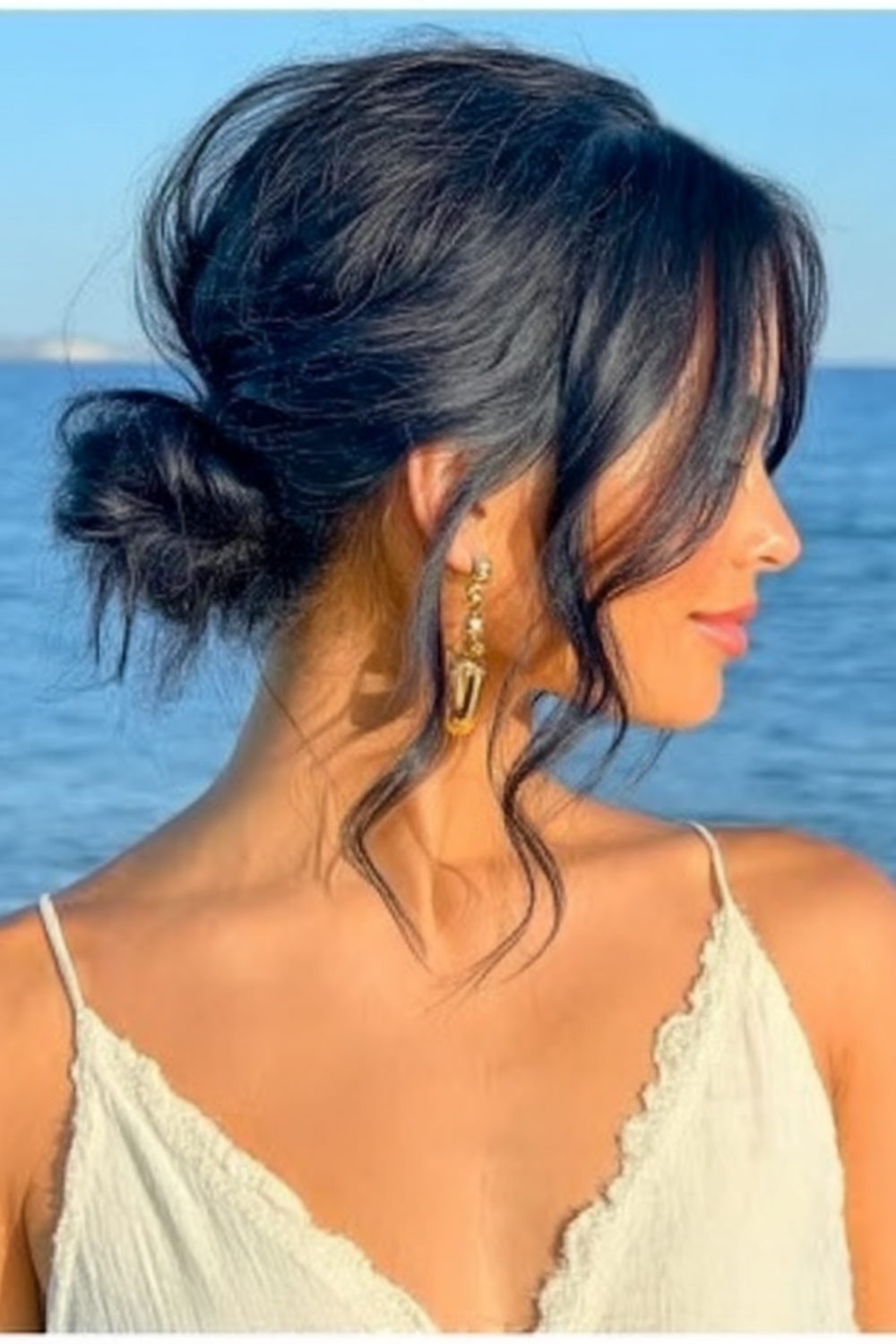 Side profile of a woman with dark hair in a loose bun, wearing a white lace-trim top and gold earrings, by the blue ocean.